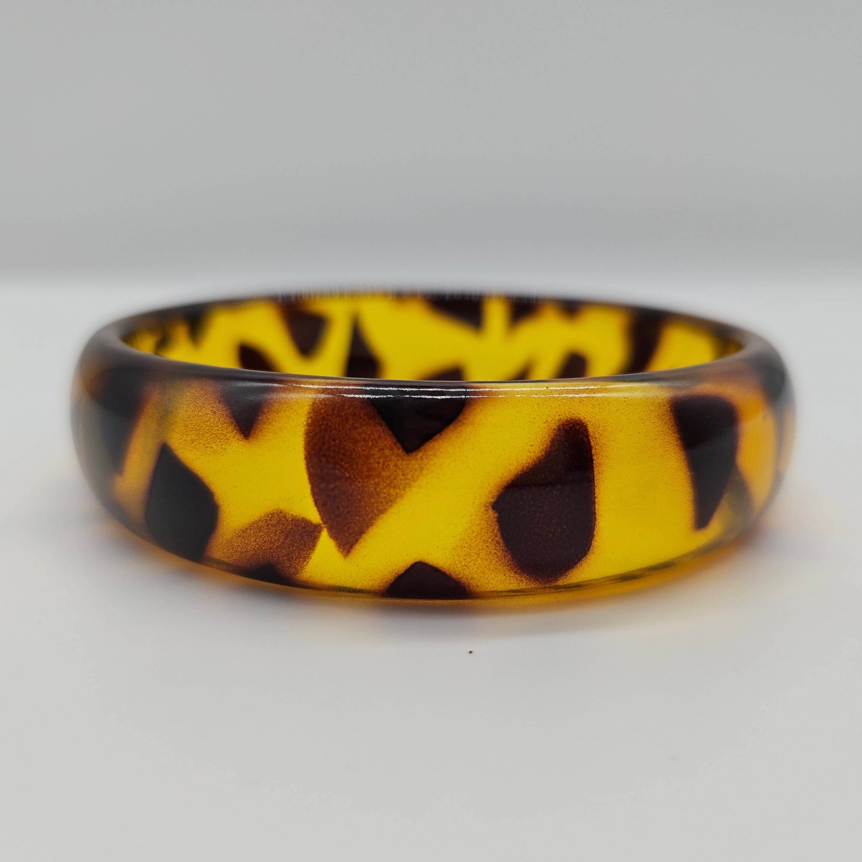 A glossy tortoiseshell-patterned bangle with a vibrant yellow base and dark brown and black mottled spots, shown on a neutral light gray background.