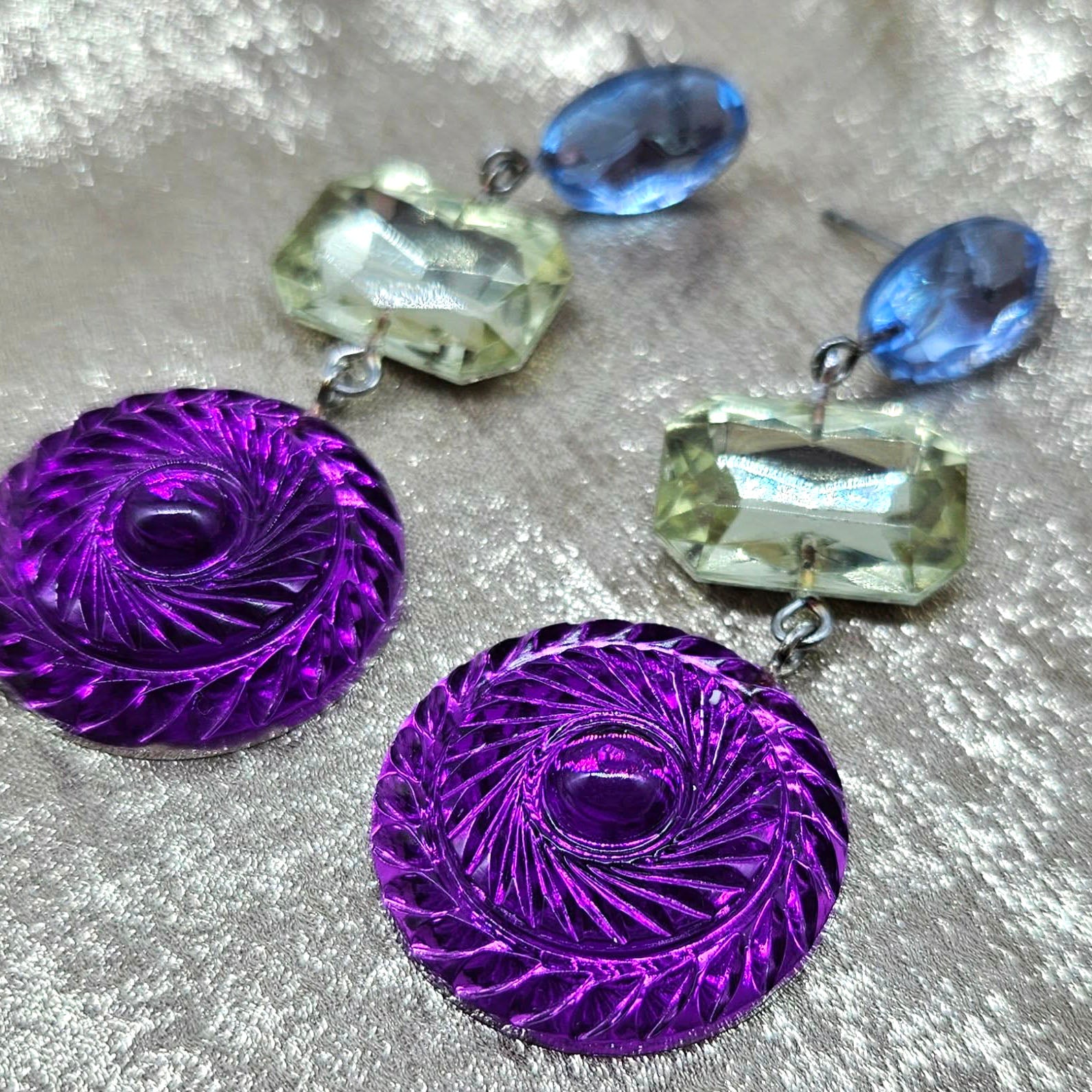 Purple circular earrings with blue and green shaped stones on a textured surface