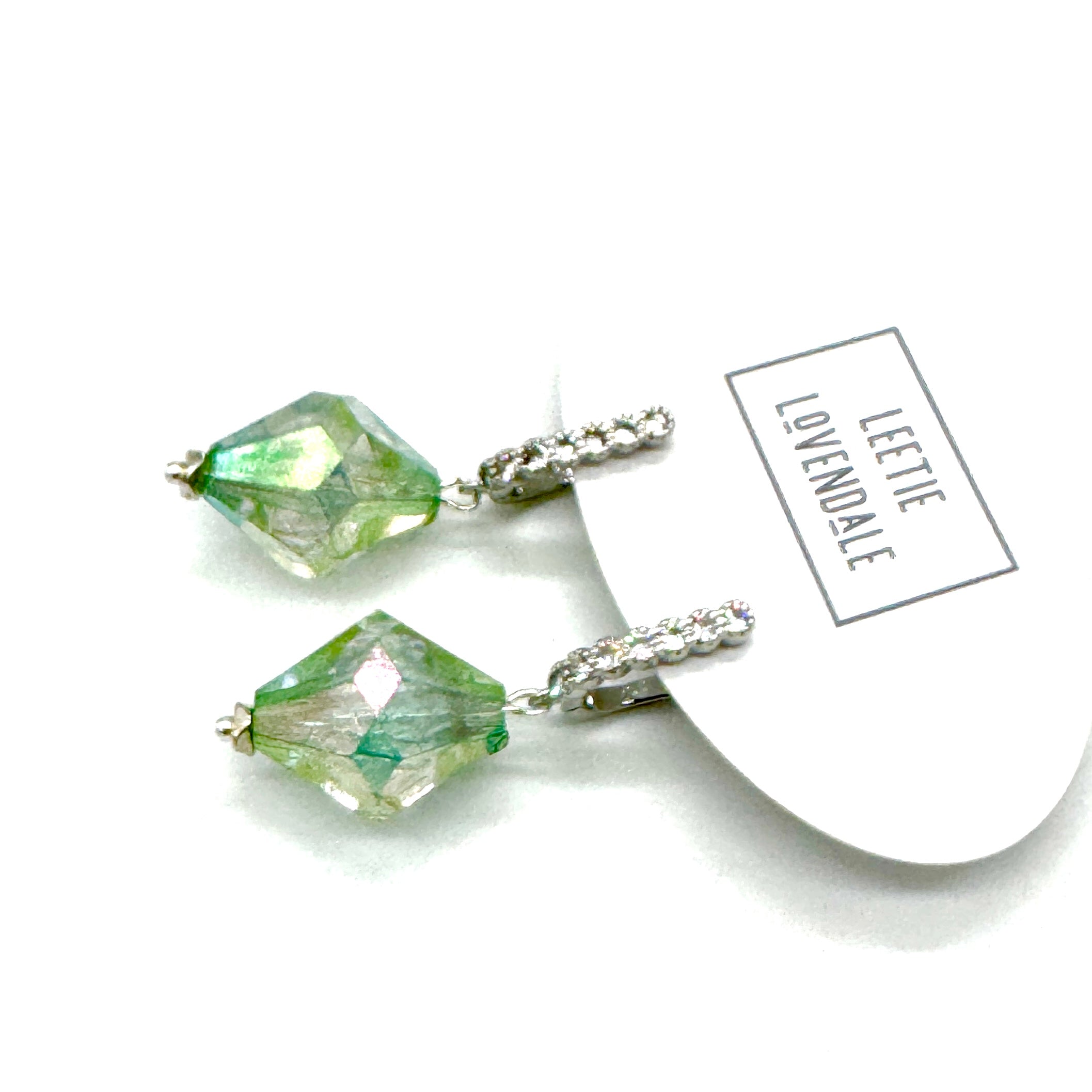 Green crystal earrings with silver hooks on a white background, featuring the brand &
