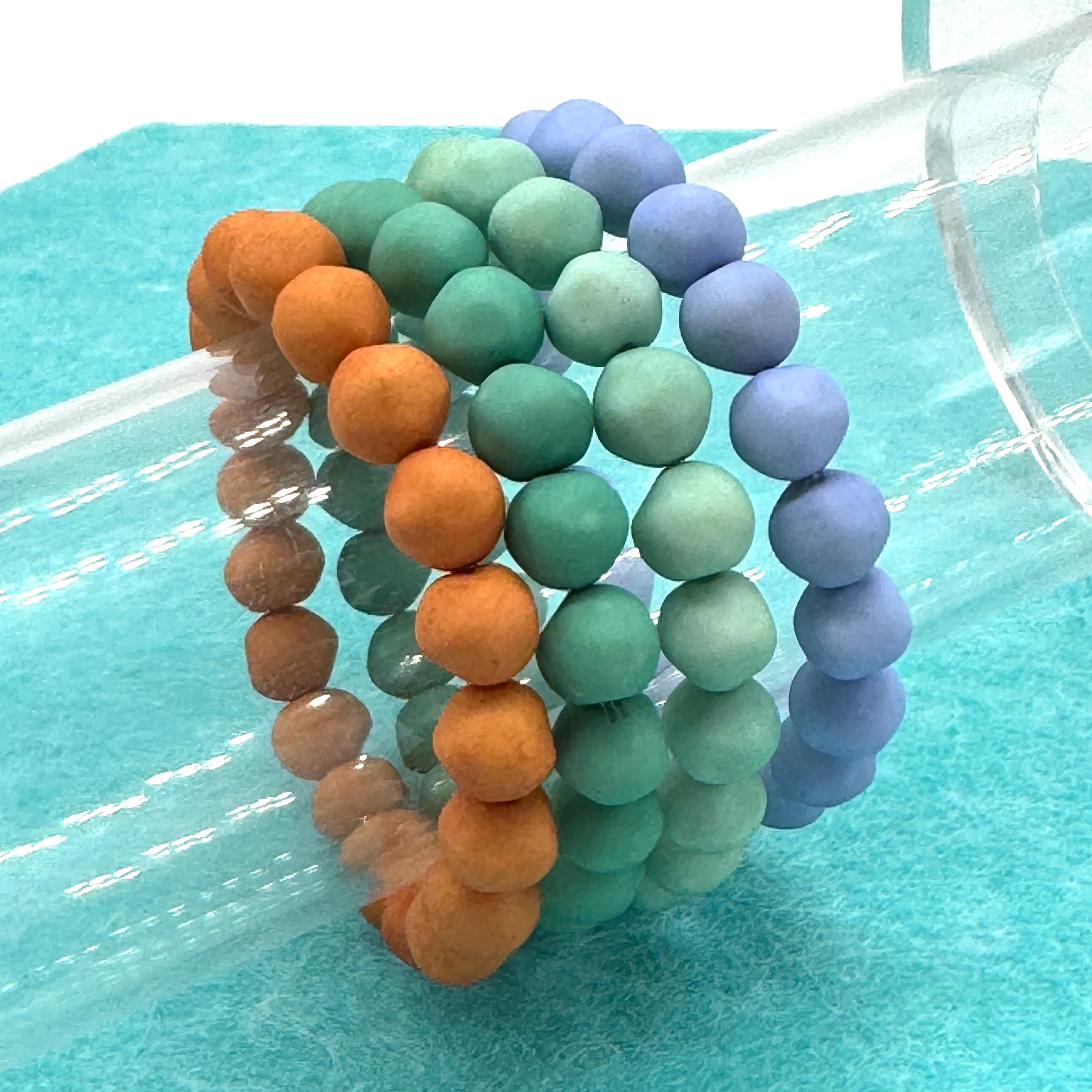 Set of four beaded bracelets in orange, green, and purple on a light blue surface.