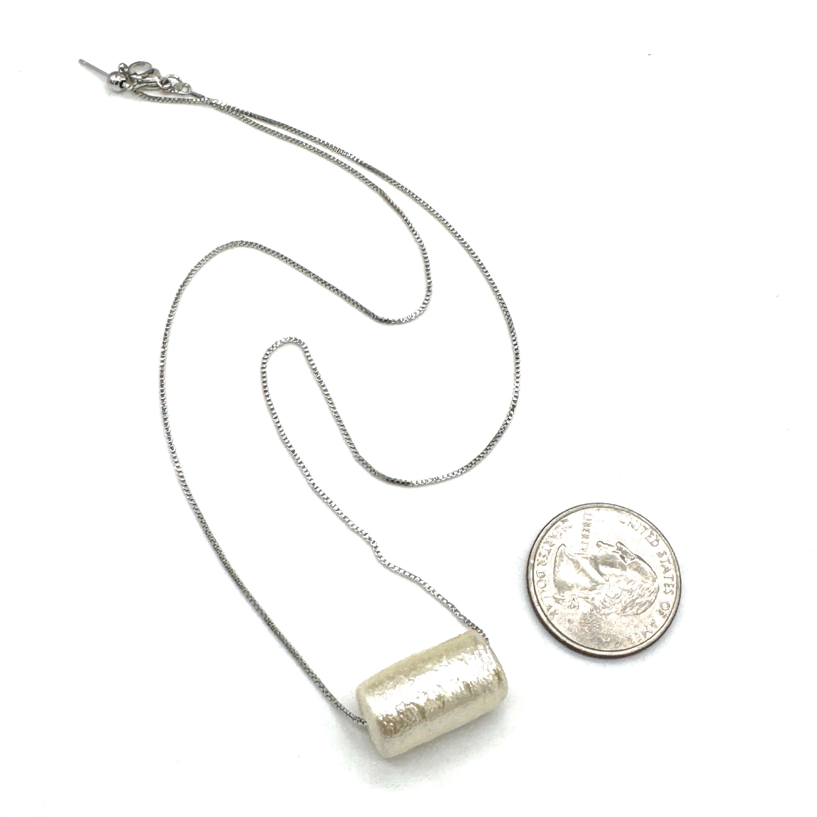 Silver necklace with a cylindrical pendant next to a quarter for scale on a white background