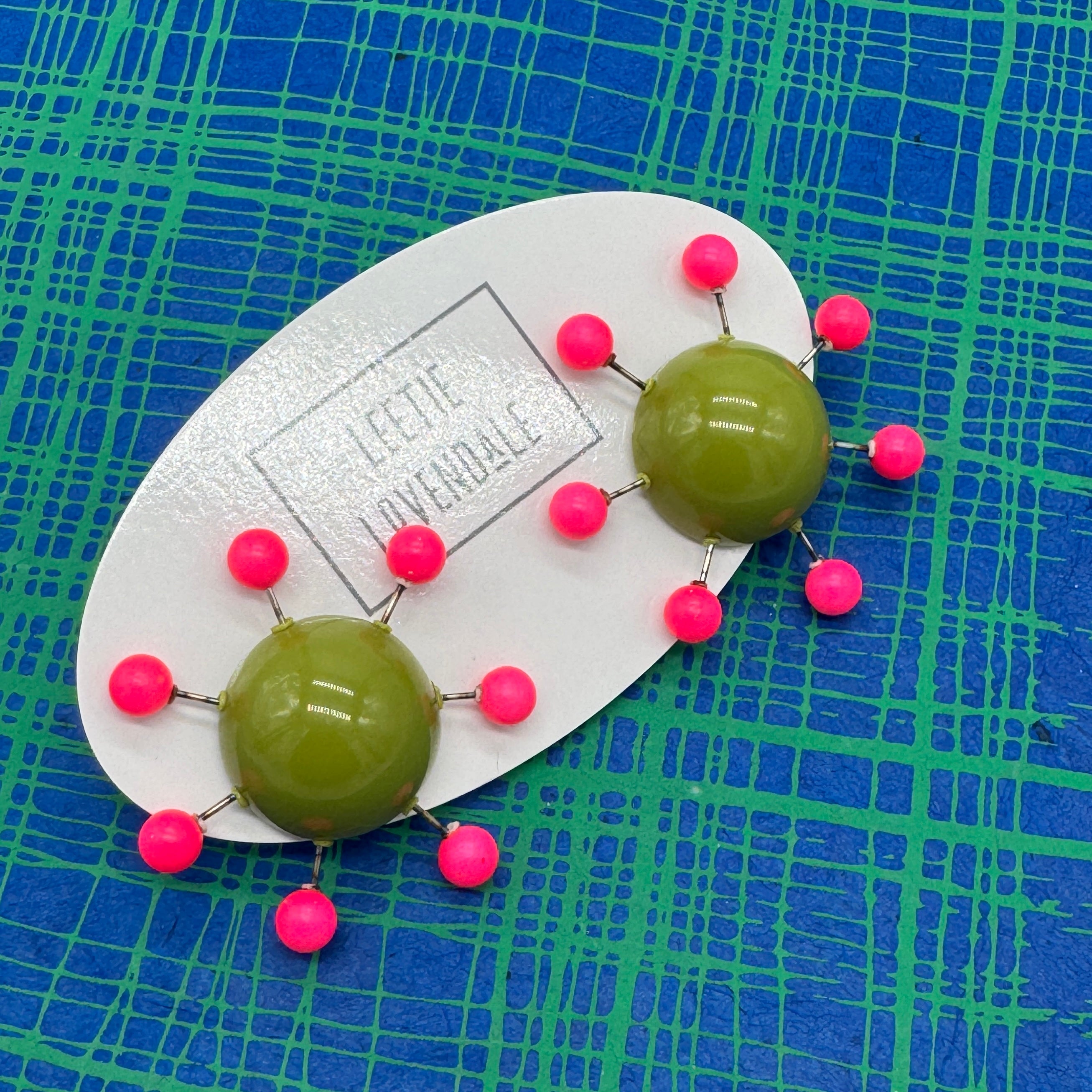 OlIve green earrings with hot pink beads radiating from center with posts on a blue and green plaid background.