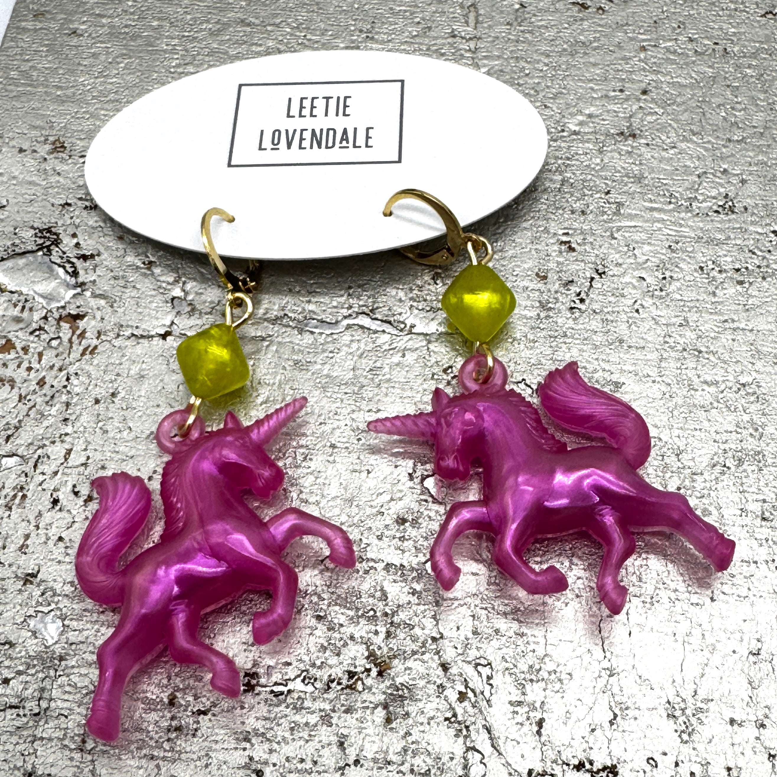 Pink unicorn earrings with green beads on a textured surface with &