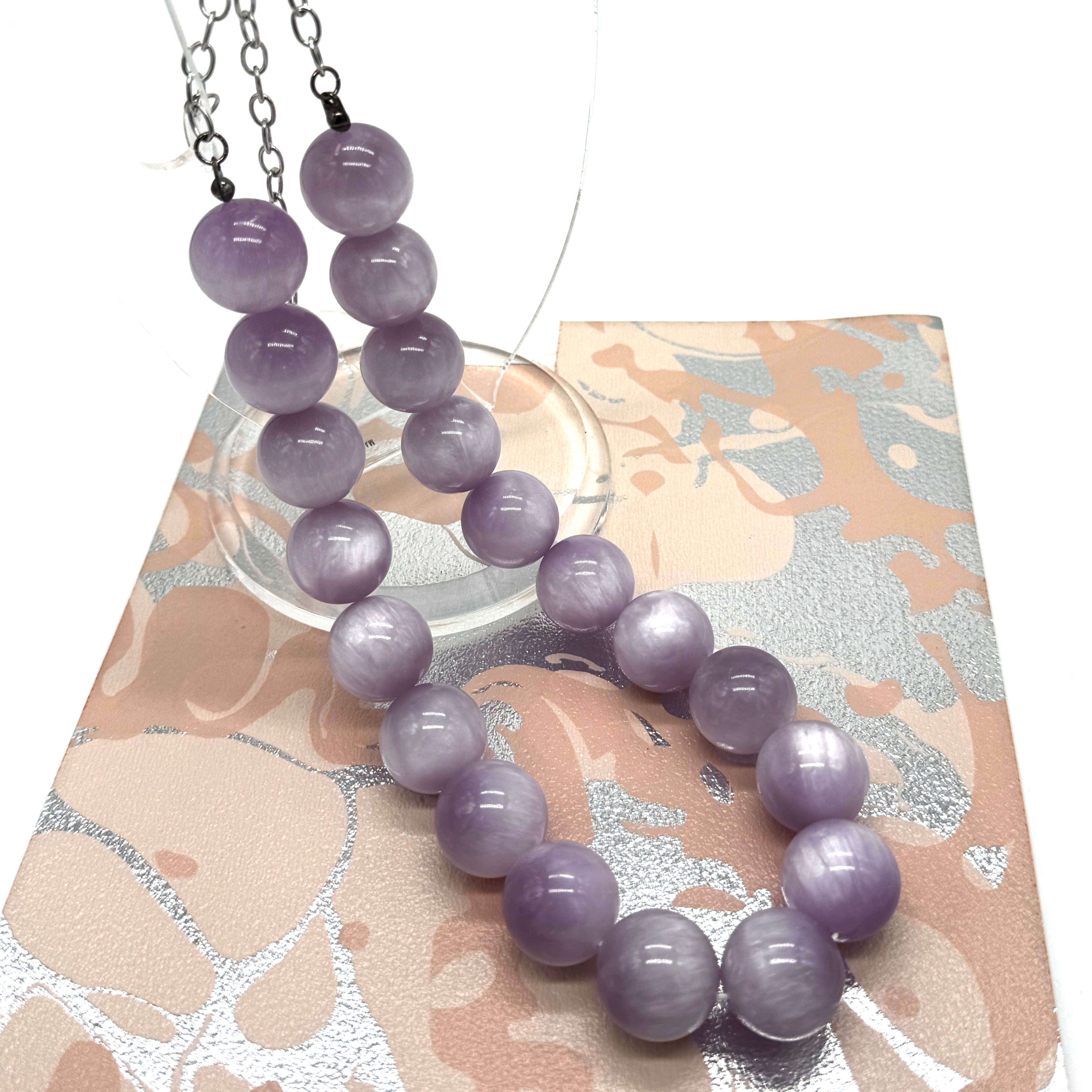 A purple round-bead necklace with silver-toned chains, displayed on a decorative beige and silver patterned card over a white background.