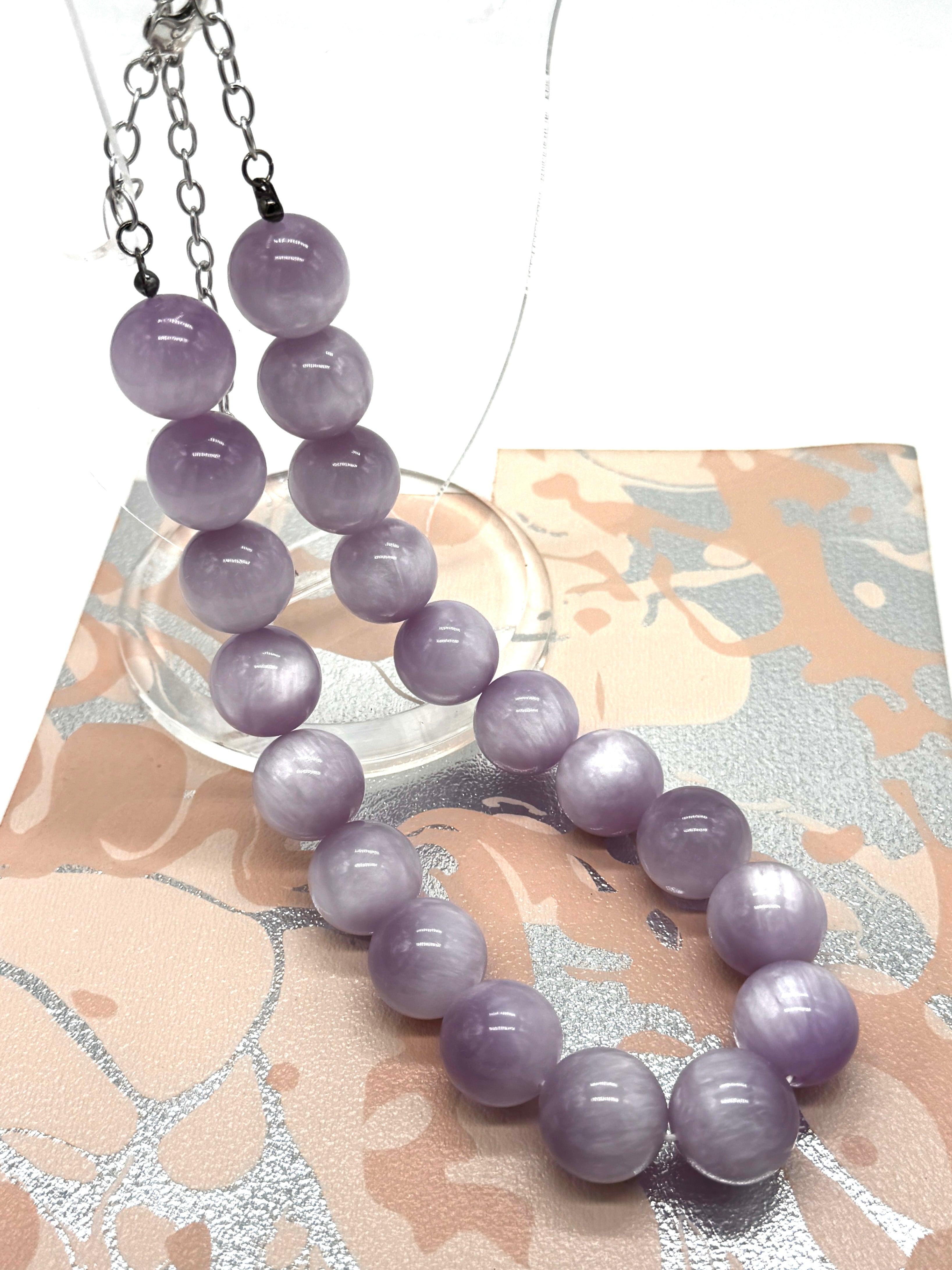 A beaded necklace made of smooth lavender-purple round beads, displayed on a patterned beige and silver card over a white background.