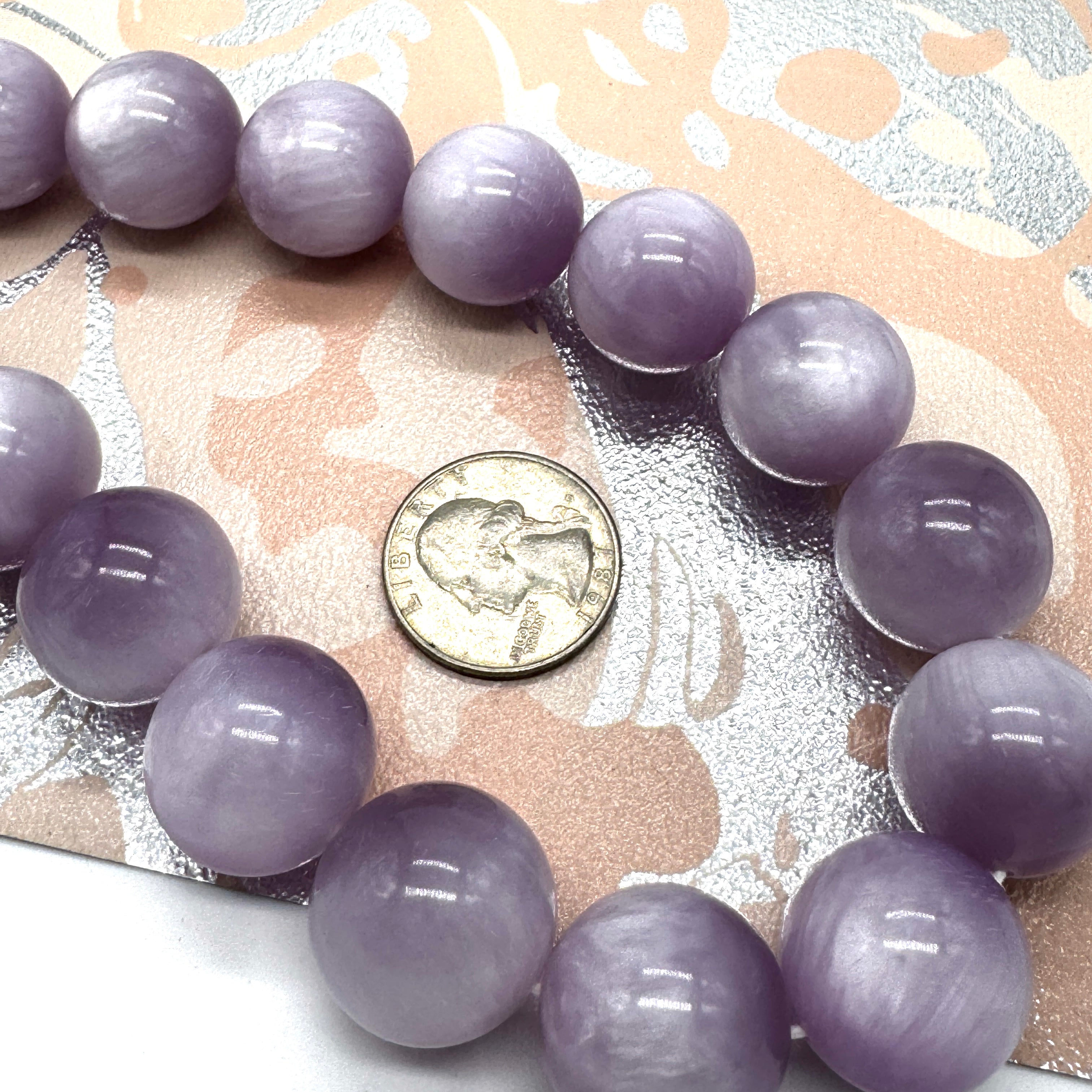 A strand of round, polished lavender-purple beads, arranged in a circle next to a US quarter coin for scale, on a patterned beige and silver background.
