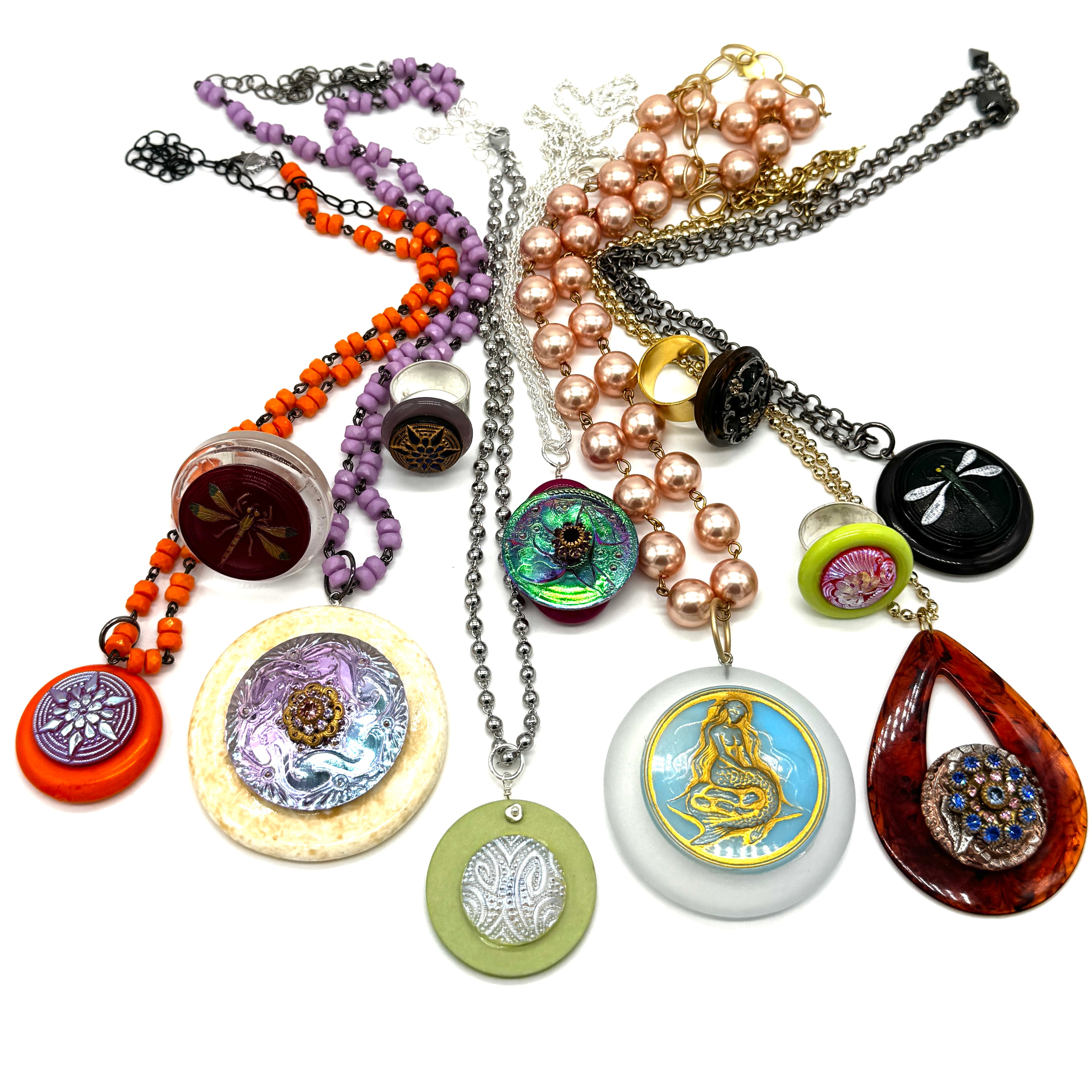 A collection of nine distinct handmade bead necklaces and pendants arranged on a plain white background. The jewelry features a variety of materials including orange beads, purple beads, faux pearls, silver chains, green and lavender beads, and colorful enamel medallions.