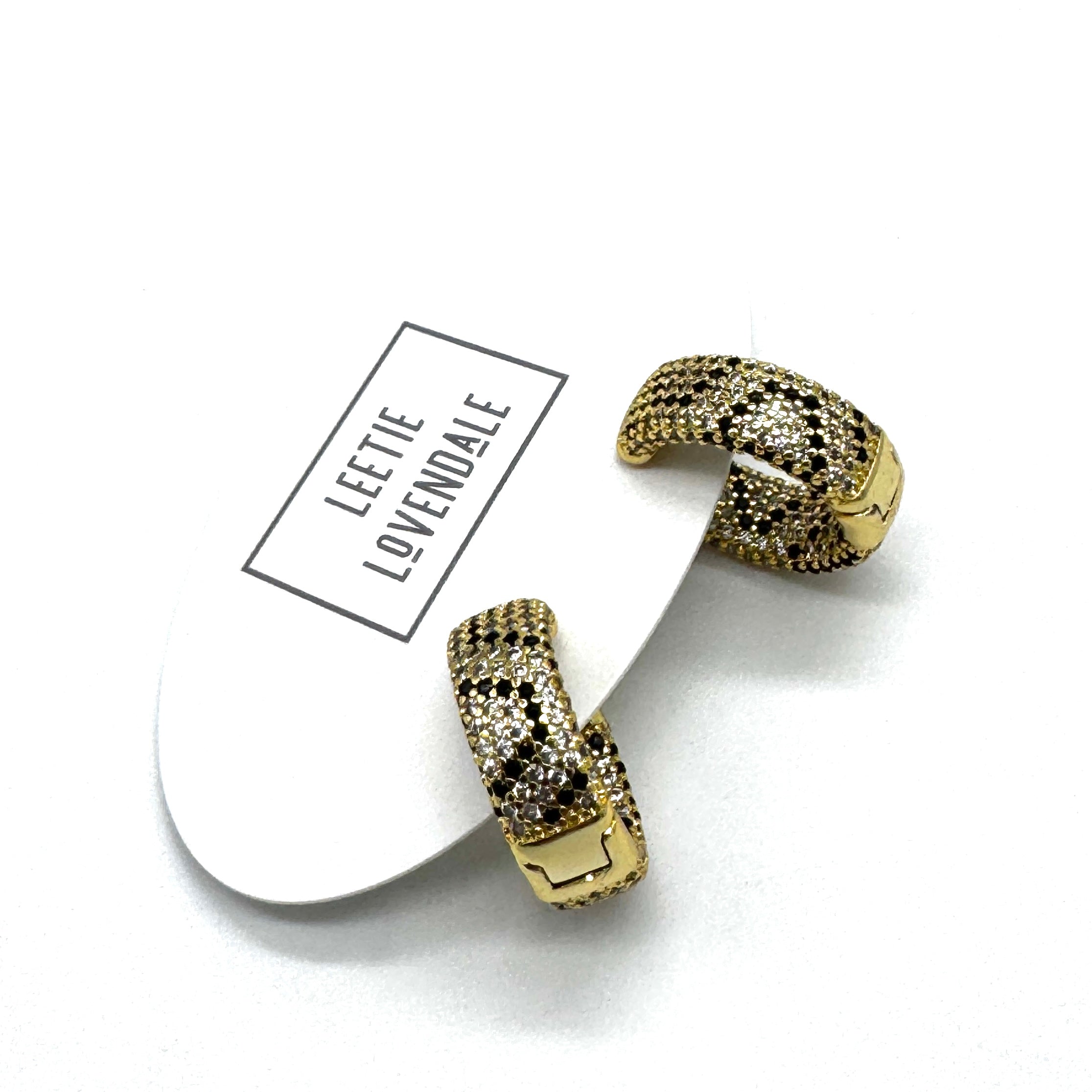 A pair of gold huggie hoop earrings covered in small black and clear rhinestones, resting on a white display card with the brand name &
