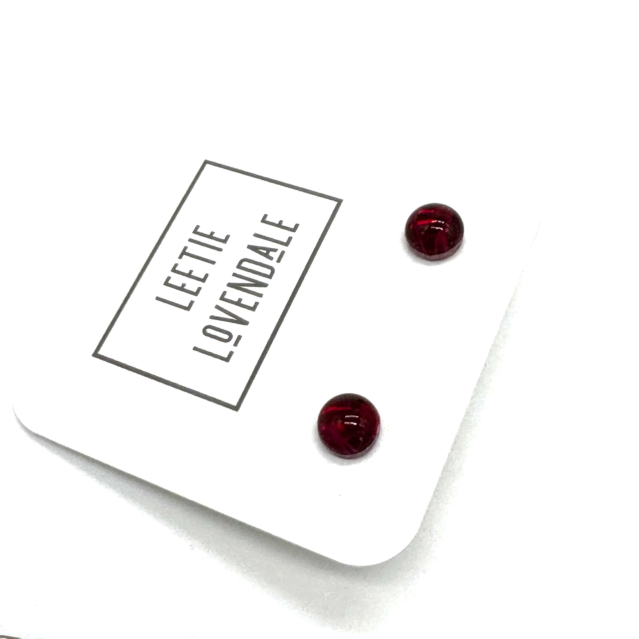 A pair of small round stud earrings in deep red resin, displayed on a white branded card that reads &