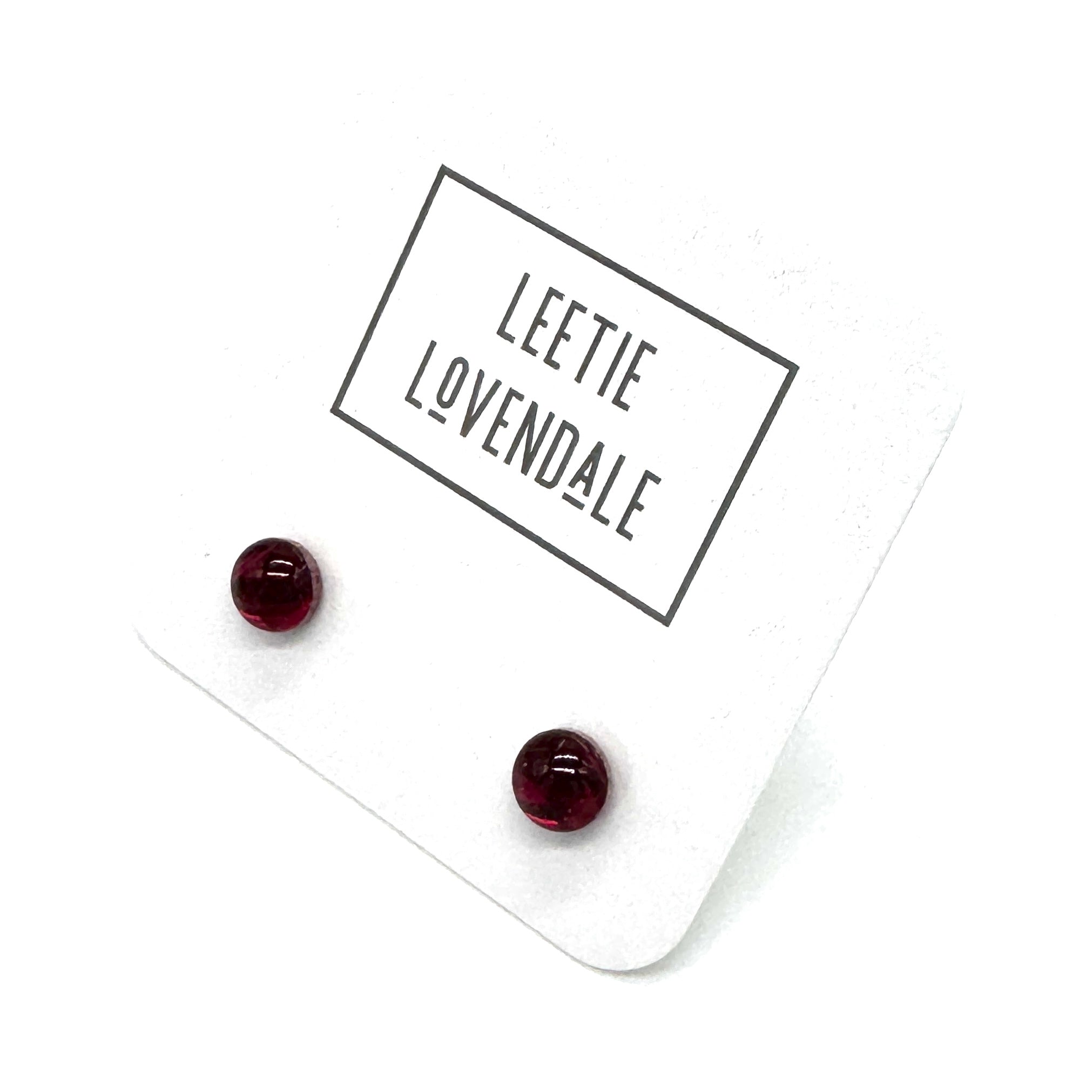 A pair of small round dark red stud earrings displayed on a white backing card with the brand name &