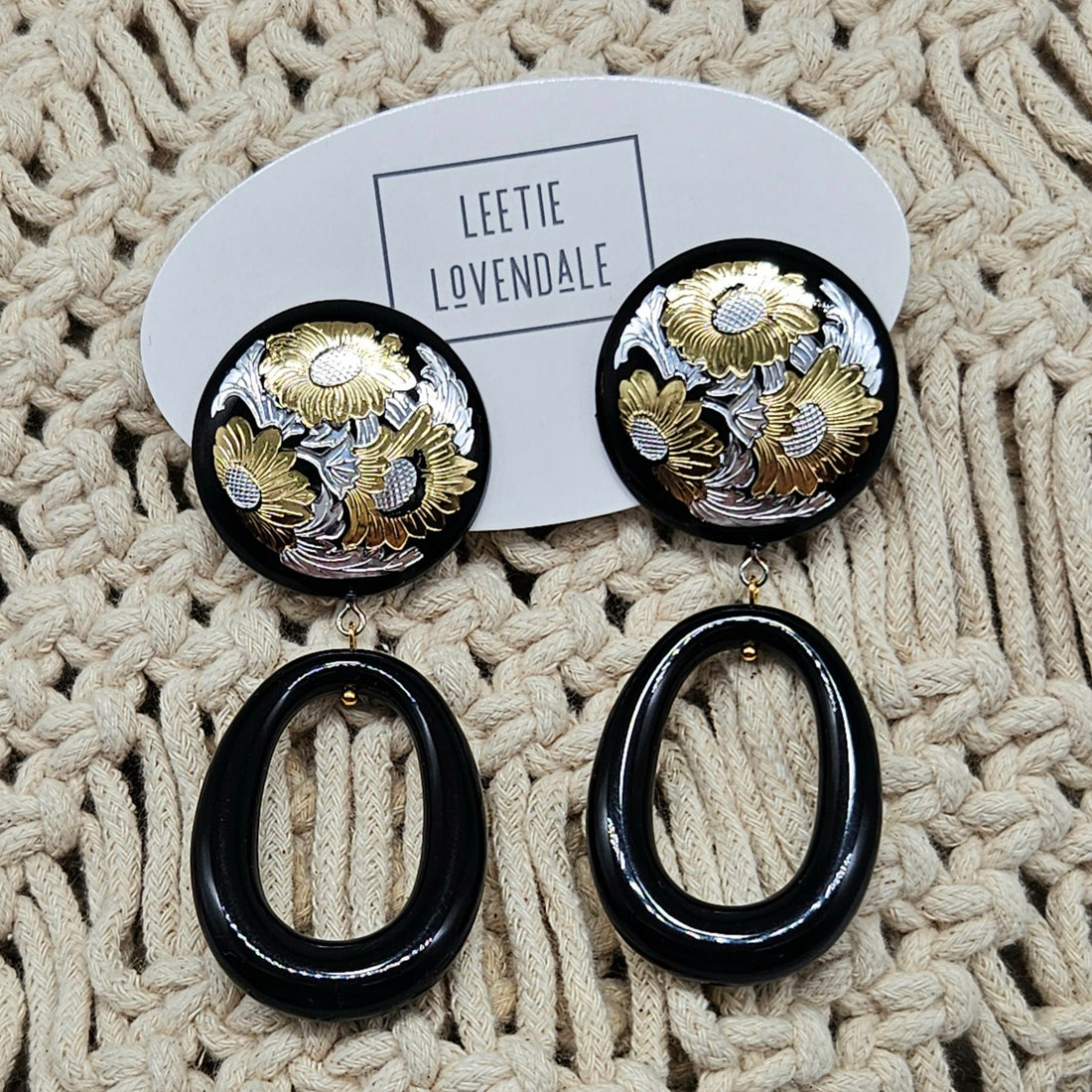 Pair of earrings with black and gold design on a textured beige surface, branded &