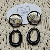 Pair of earrings with black and gold design on a textured beige surface, branded &