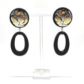 Pair of earrings with floral design and black oval shapes on a clear stand.