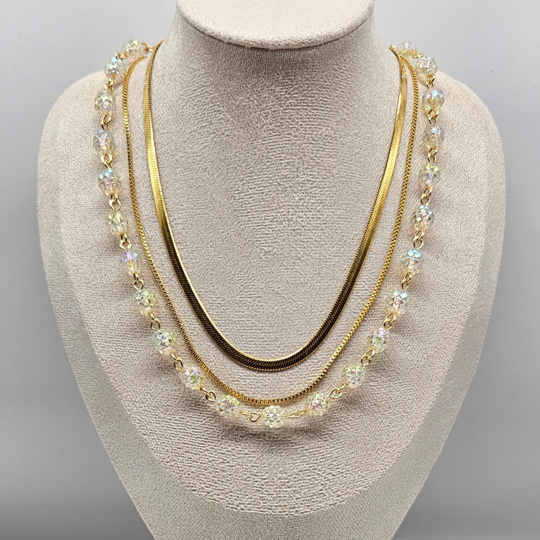 Three-strand gold necklace with clear lucite beads and chains on a mannequin bust against a gray background