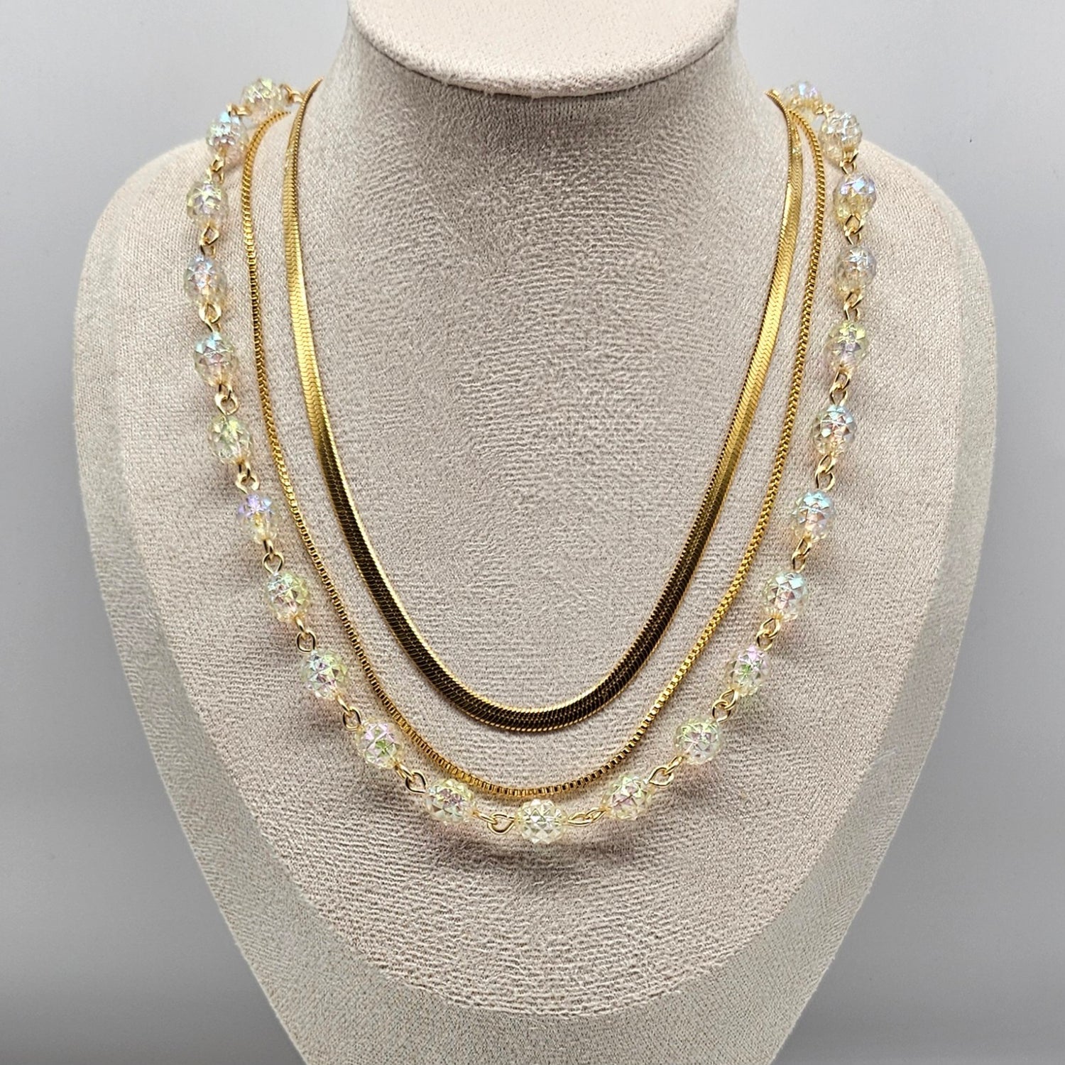 Three-strand gold necklace with clear lucite beads and chains on a mannequin bust against a gray background