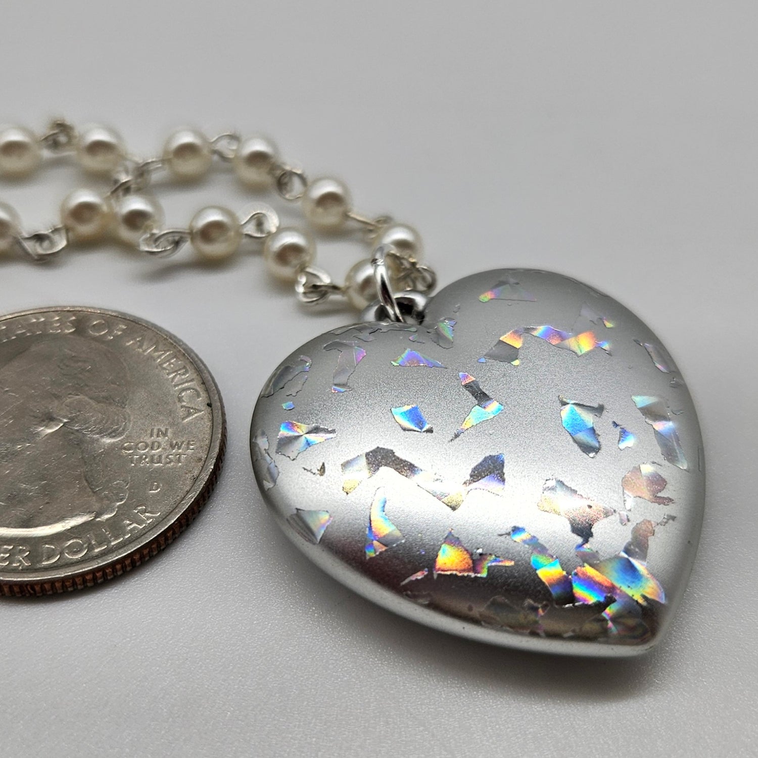 Heart-shaped silver pendant with colorful inclusions next to a quarter on a gray background
