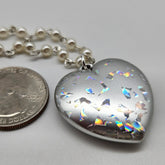 Heart-shaped silver pendant with colorful inclusions next to a quarter on a gray background