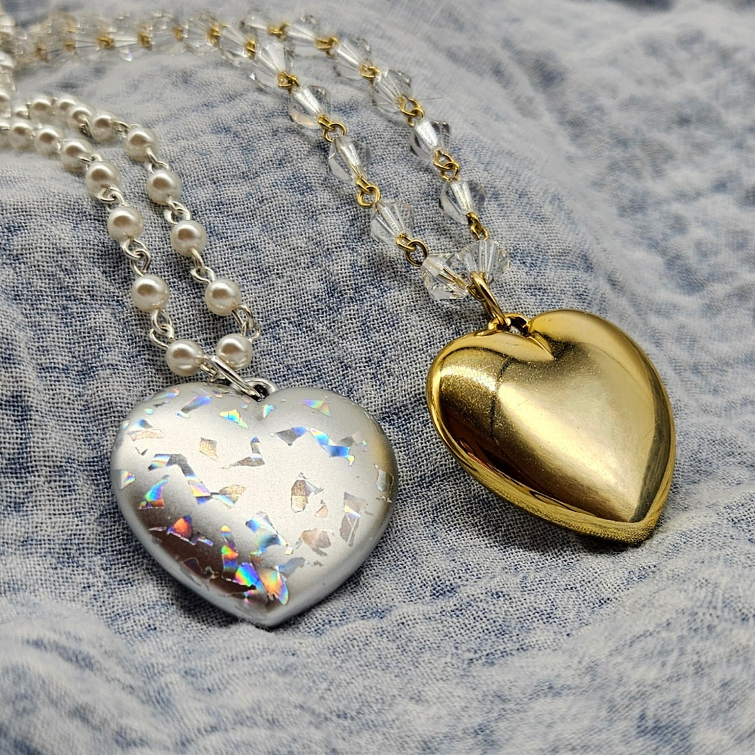 Two heart-shaped pendants, one silver and one gold, on a textured fabric background.