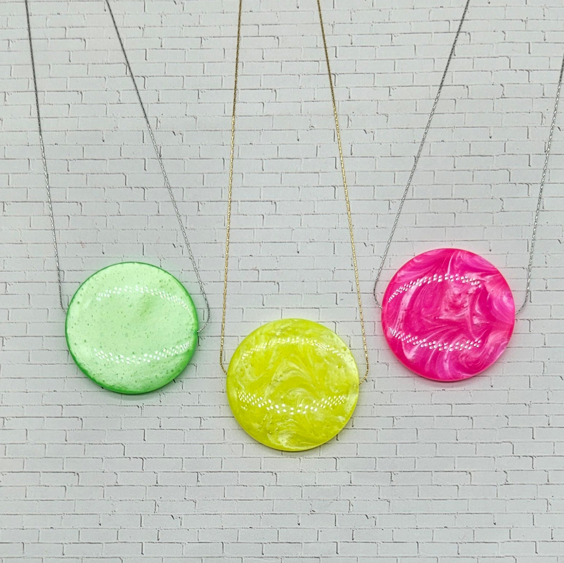 Three colorful circular pendants on necklaces against a white brick wall background.