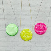 Three colorful circular pendants on necklaces against a white brick wall background.