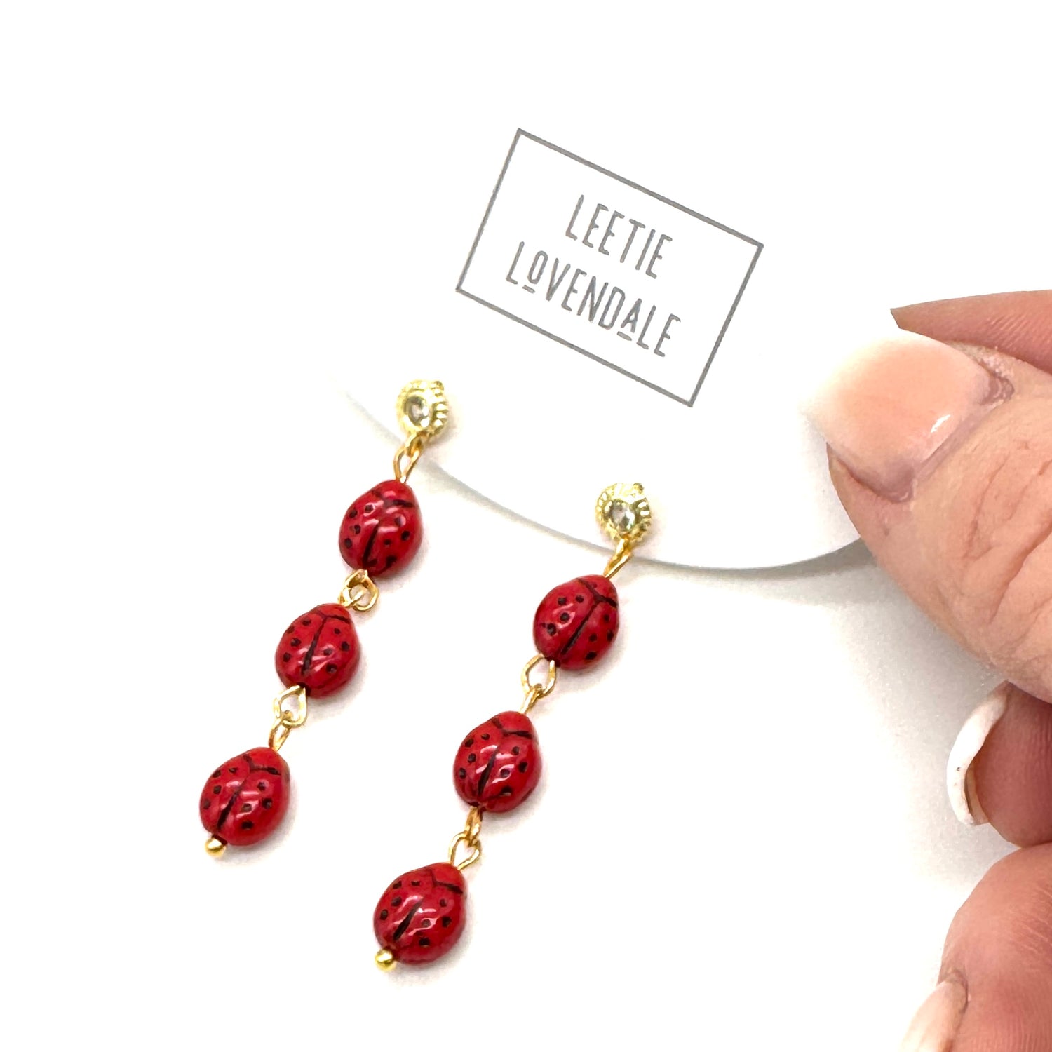 Red beaded earrings with gold accents held by a hand, brand &