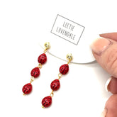 Red beaded earrings with gold accents held by a hand, brand &