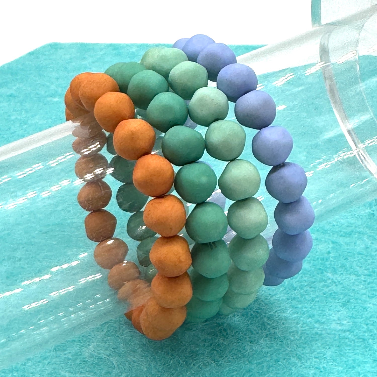 Set of four beaded bracelets in orange, green, and purple on a light blue surface.