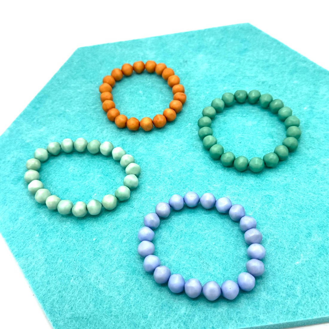 Four beaded bracelets in orange, green, and purple on a turquoise hexagonal mat.