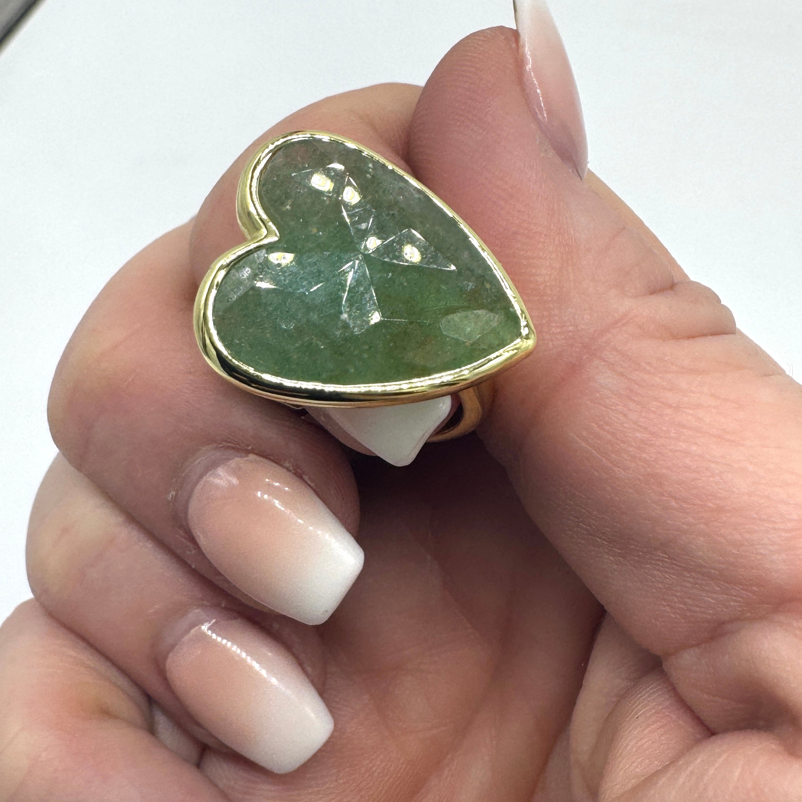 Heart-shaped green stone ring with gold border on a hand against a white background