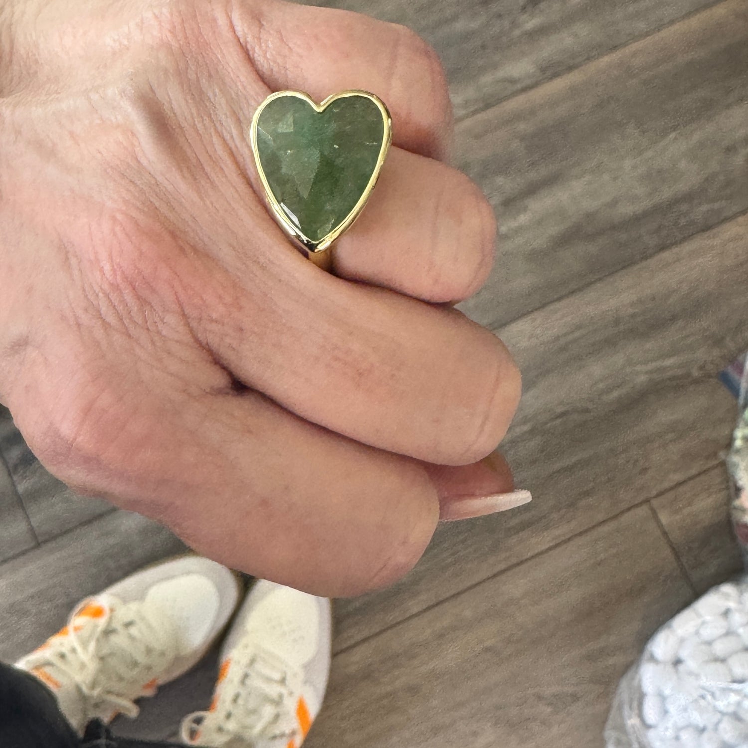 Hand wearing a heart-shaped green stone ring on a wooden floor.