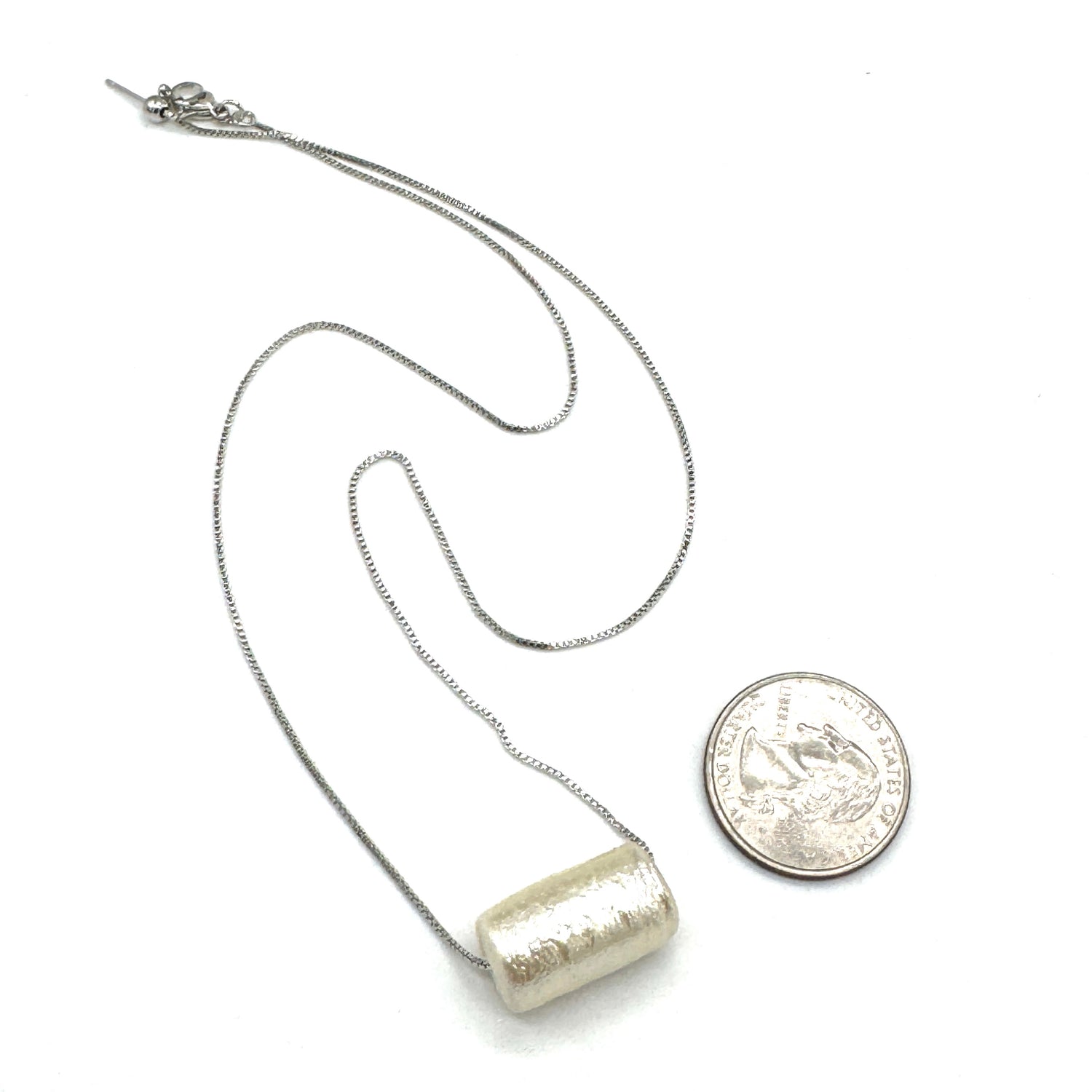 Silver necklace with a cylindrical pendant next to a quarter for scale on a white background