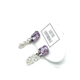 Pair of purple crystal earrings with silver chains on a white background, featuring a &