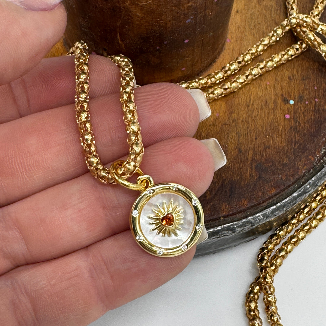 Gold necklace with a sun  and heart pendant held by a hand on a wooden surface
