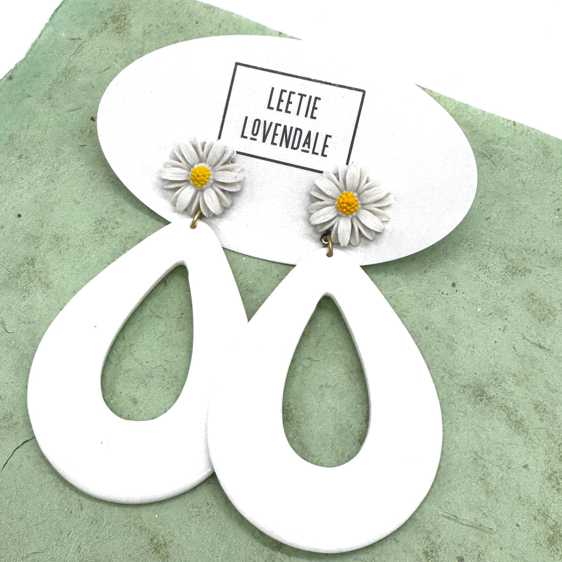 White earrings with floral designs on a green surface, featuring the brand &