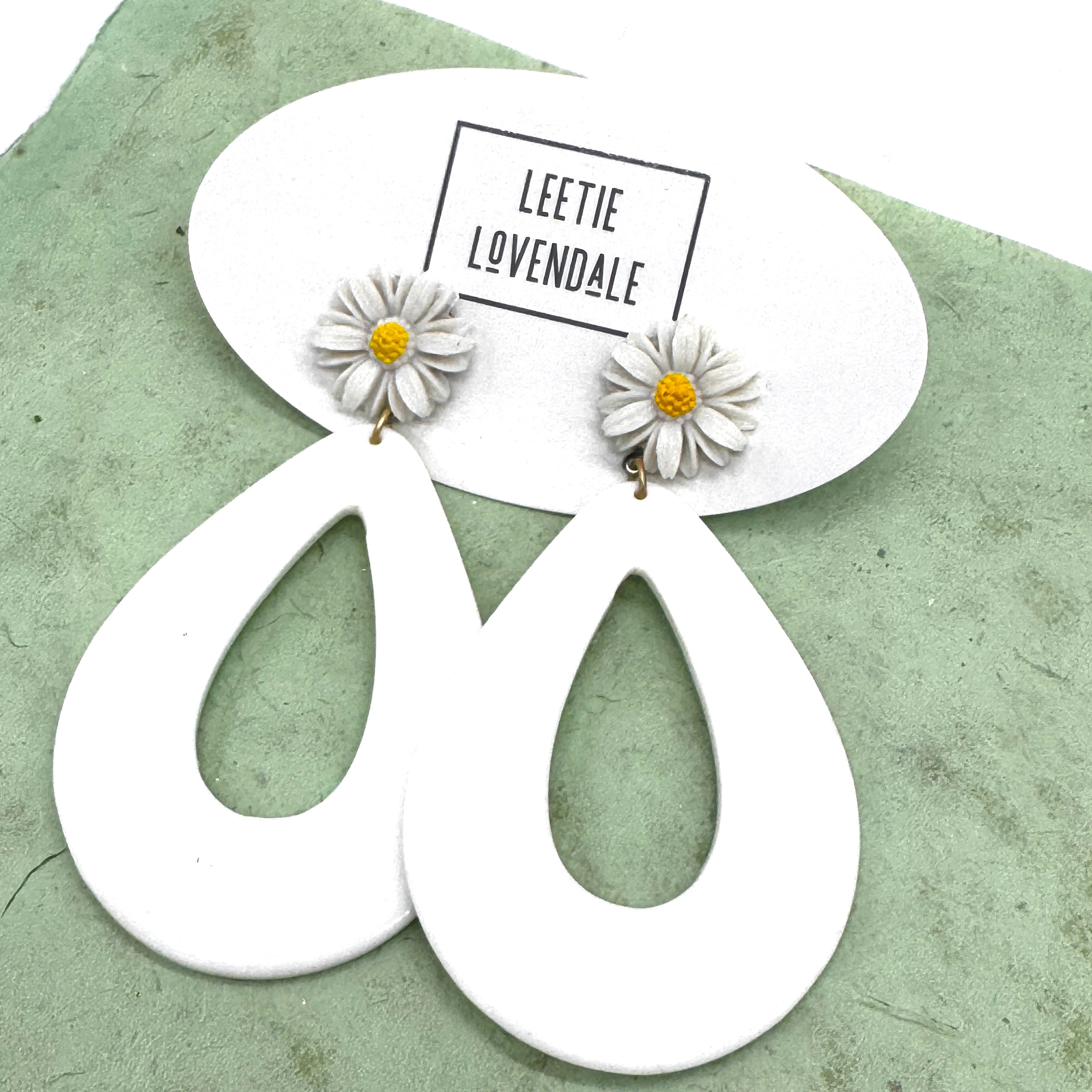 White earrings with floral designs on a green surface, featuring the brand &