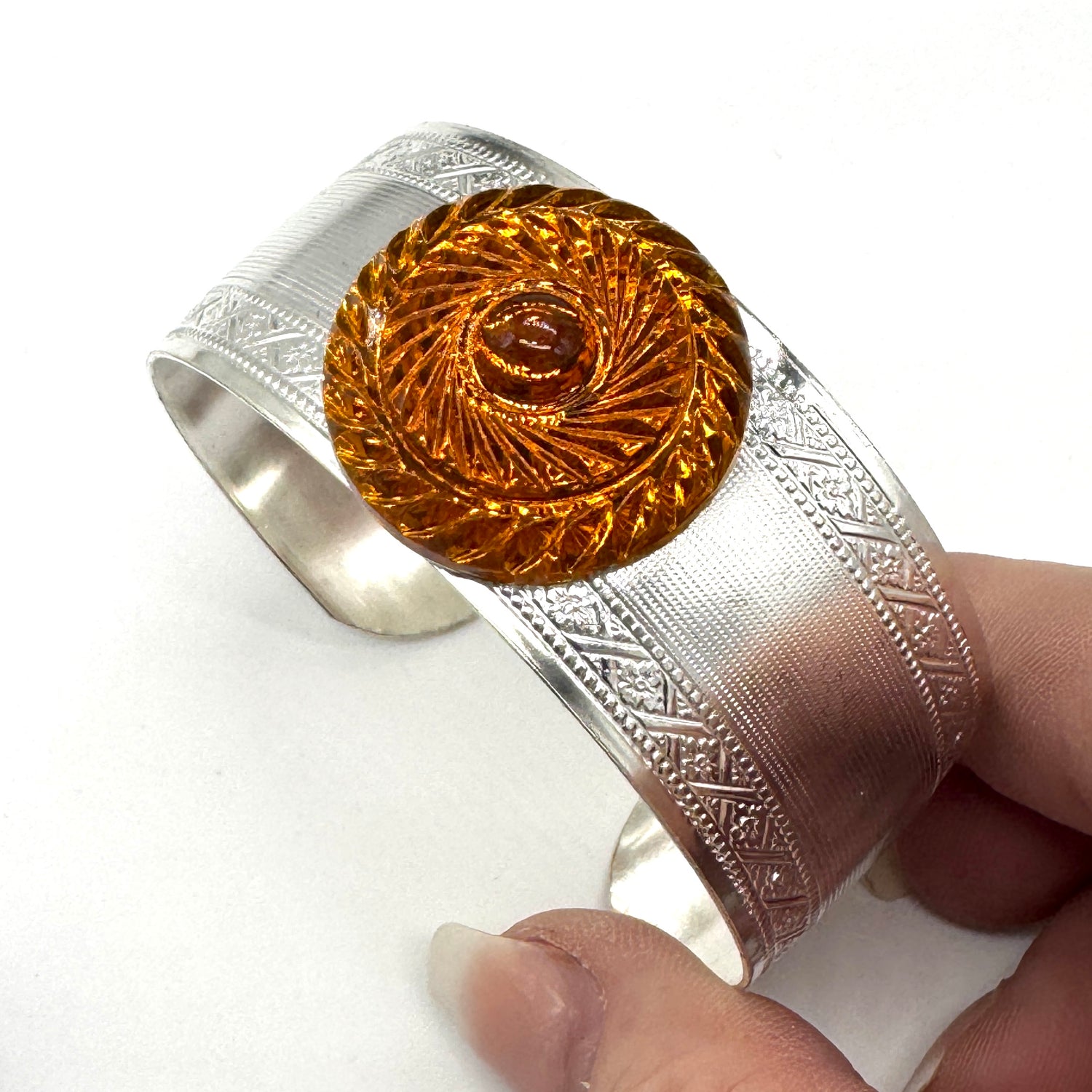 Silver bracelet with an orange lucite stone with a circular design held by a hand on a white background