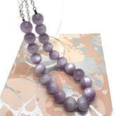 A purple round-bead necklace with silver-toned chains, displayed on a decorative beige and silver patterned card over a white background.