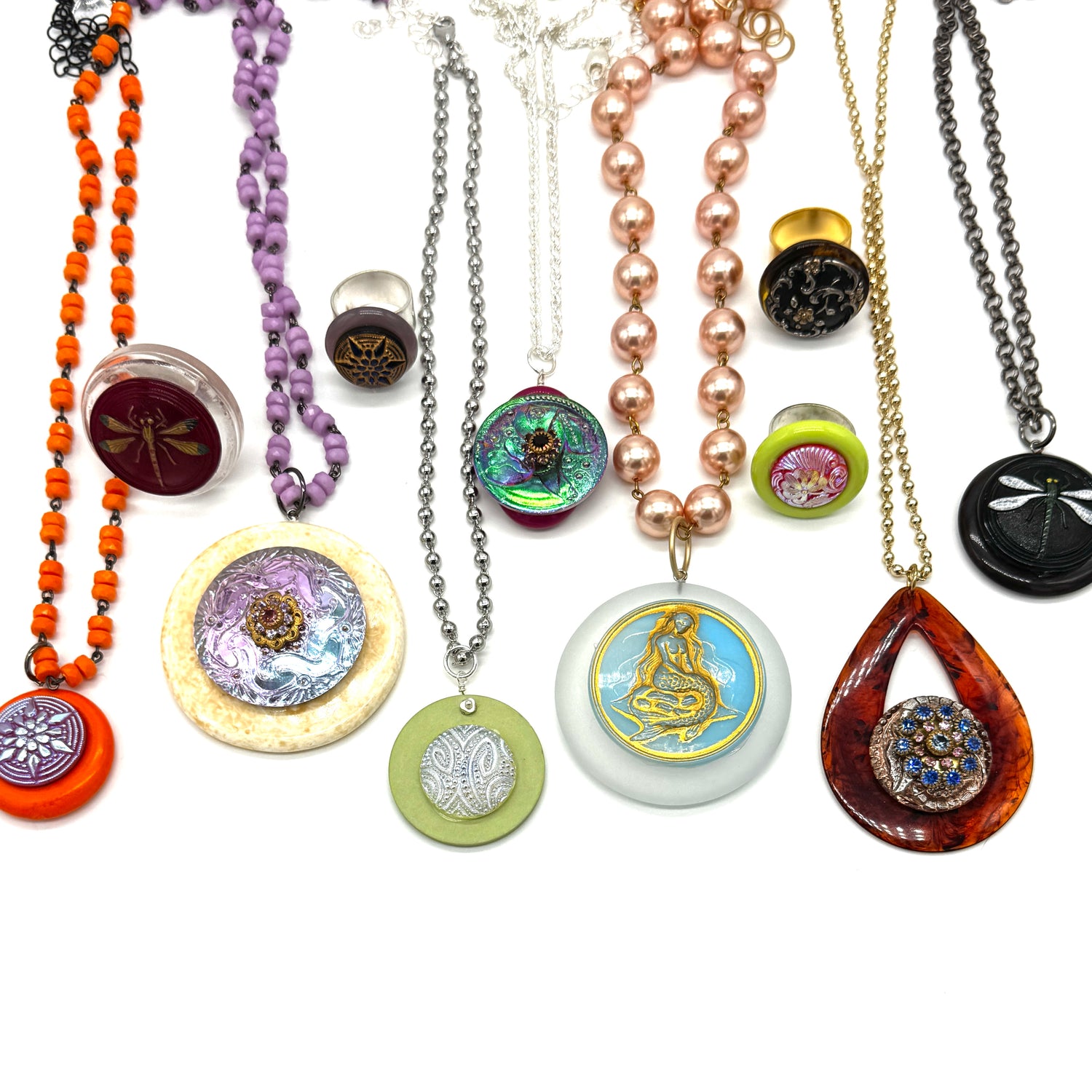 A collection of handmade bohemian-style necklaces and earrings displayed on a white background. The jewelry features diverse materials including orange beads, purple beads, rose gold beads, silver chains, green and lavender stone pendants, and dragonfly motifs.