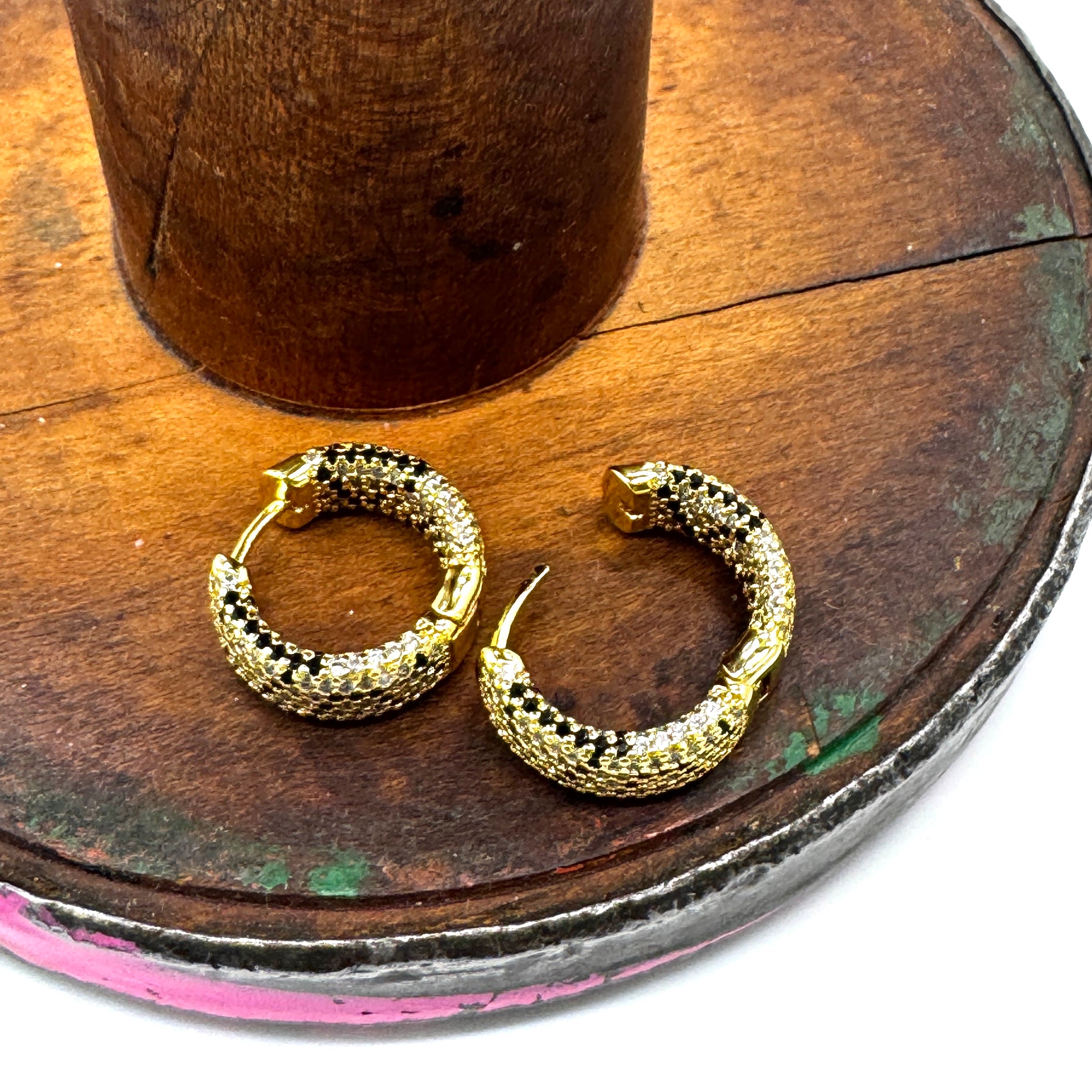 A pair of gold hoop earrings with a textured, snake-like pattern, displayed on a rustic wooden stand.