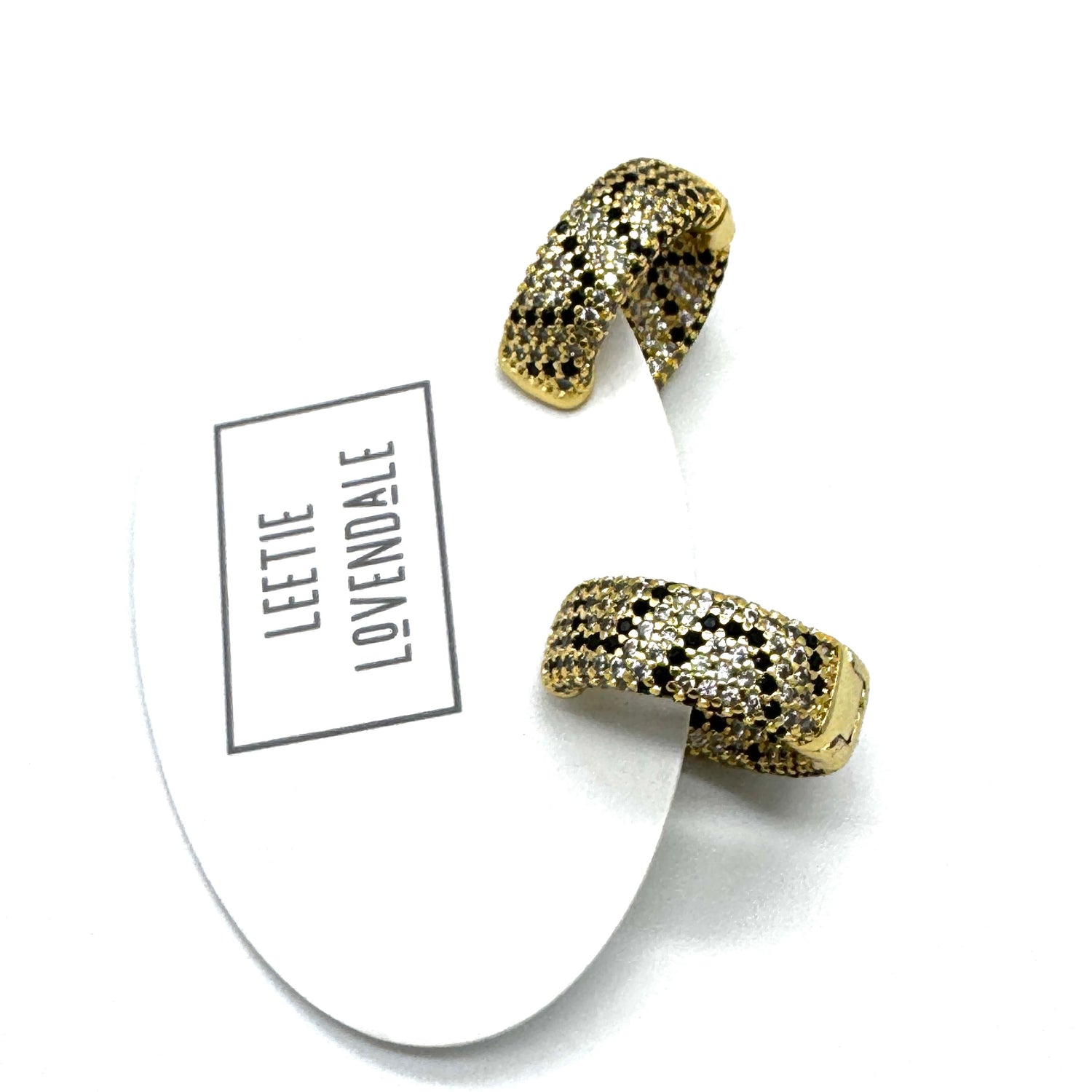 A pair of gold huggie hoop earrings covered in small black and white stones, displayed on a white branded card that reads &