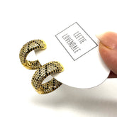 A pair of gold hoop earrings covered in small black and clear rhinestones, attached to a white display card held by a hand. The card is branded &