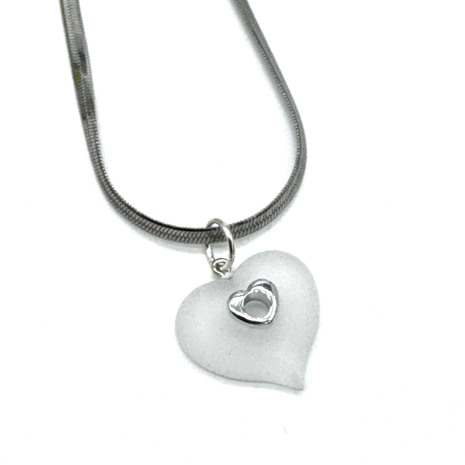 A silver snake chain necklace featuring a frosted white heart-shaped pendant with a small, polished silver heart inset in the center, shown against a plain white background.