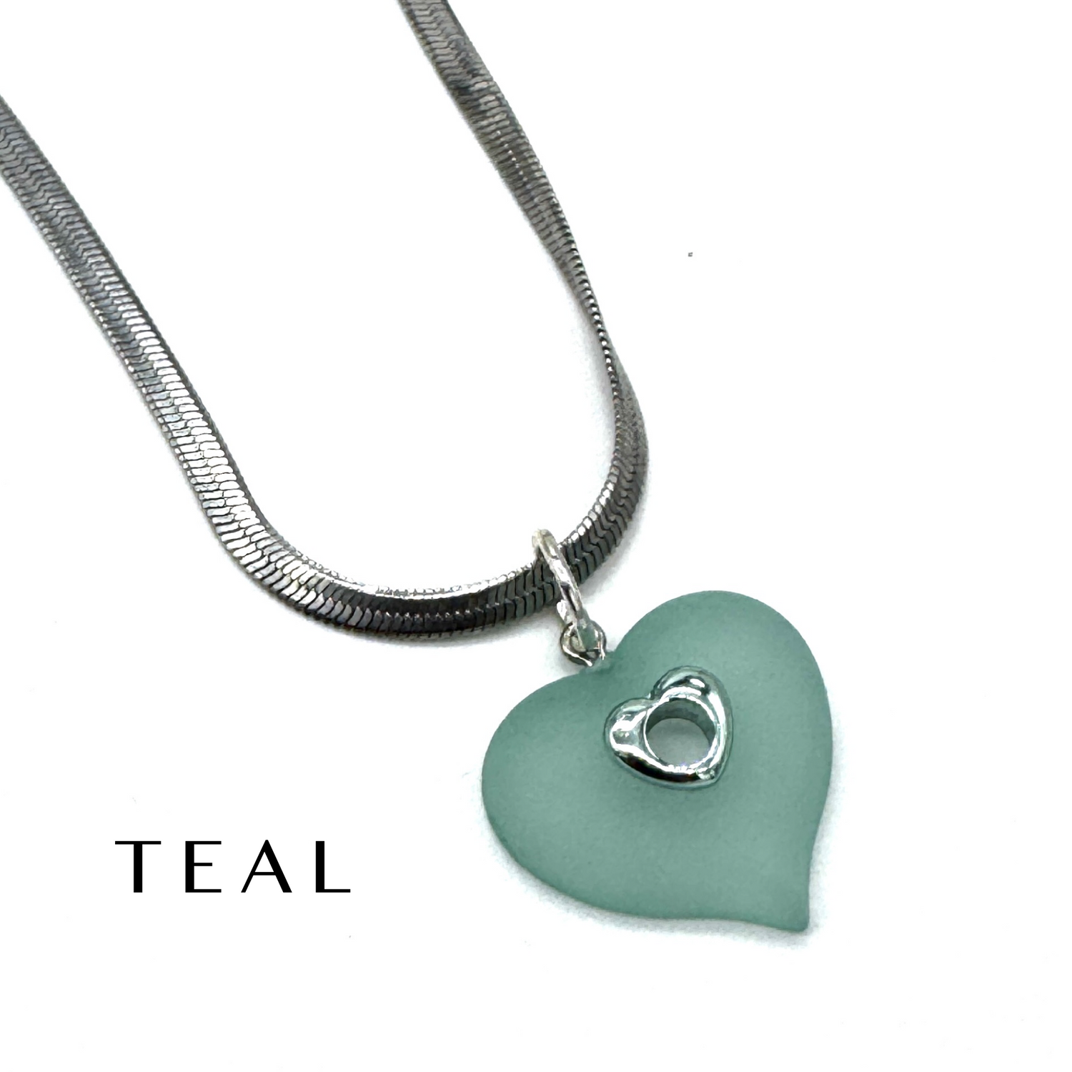 A teal heart-shaped pendant necklace with a silver snake chain and a small silver heart detail, shown on a white background with the word &