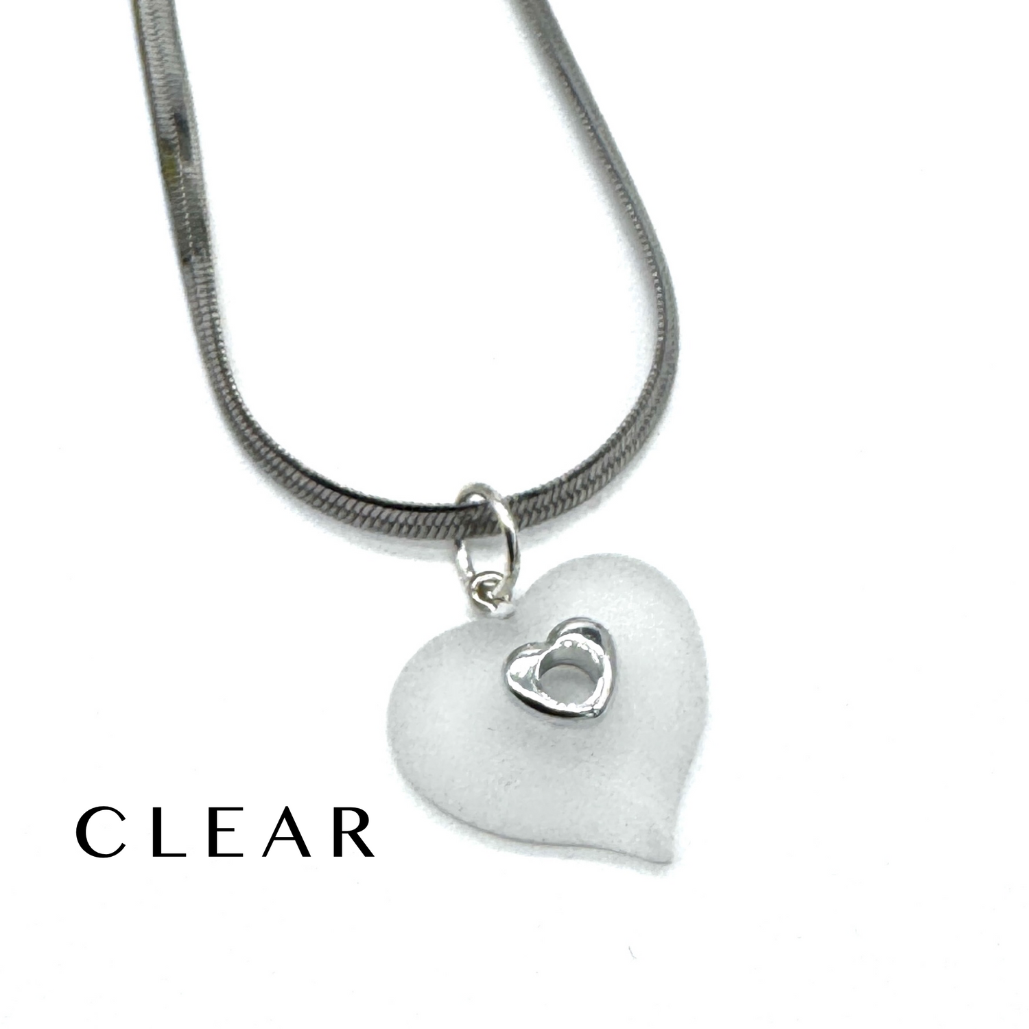 A silver snake chain necklace with a frosted clear heart-shaped pendant featuring a small silver heart in the center, shown on a white background with the word &