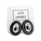A pair of oval stud earrings with shiny silver-toned centers and dark textured outer borders, displayed on a white branded backing card that reads LEETIE LOVENDALE.