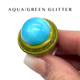 Aqua and green glittery ring held by a hand against a white background