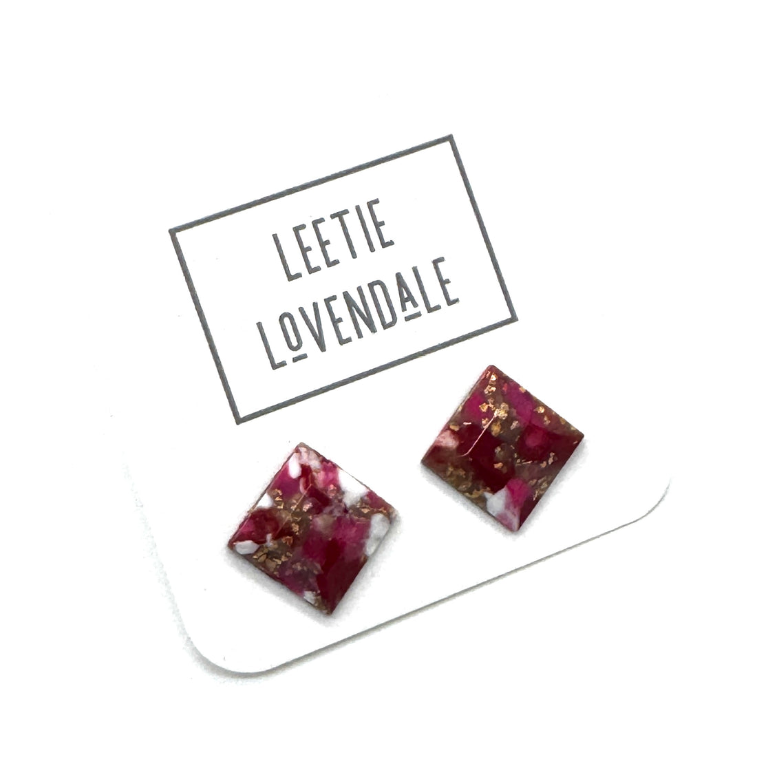 A pair of square stud earrings with a deep magenta-pink and white marbled pattern and embedded gold flakes, displayed on a white card printed with the brand name &