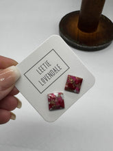 A hand holds a white display card featuring two small square stud earrings with a pink, red, and white marbled pattern and gold foil flecks. The card is branded &