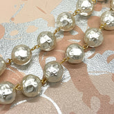 A close-up of a pearl-like beaded chain with hammered, textured round beads connected by small gold-tone links, laid diagonally across a patterned surface with pink, beige, and silver tones.