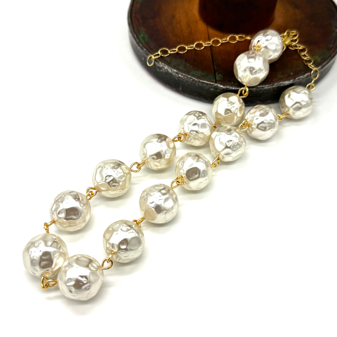 A strand necklace made of irregular, hammered white pearls connected by delicate gold chains, displayed on a clean white background with a dark wooden stand partially visible in the upper right.